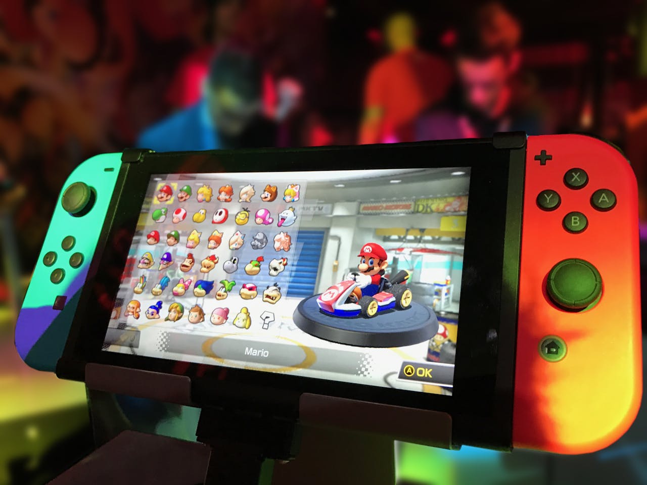 services-03 Close-up of a Nintendo Switch showing Mario Kart selection screen, gaming atmosphere in the background.